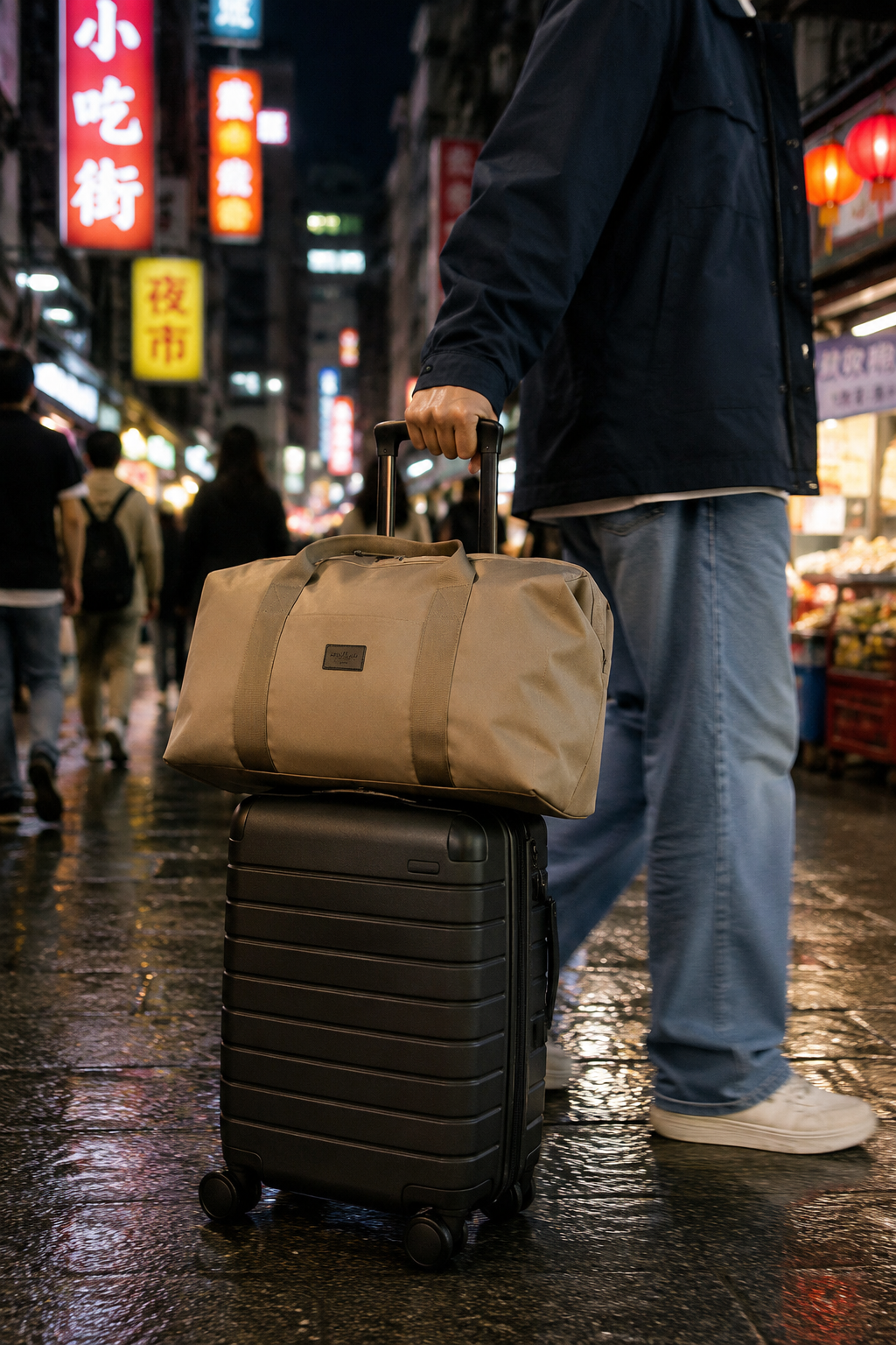 Person with a roamio duffel bag and black suitcase on a wet street at night with illuminated signs in the background
